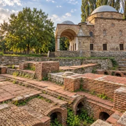 Тухлени археологически останки пред историческа каменна сграда с купол, заобиколена от дървета.