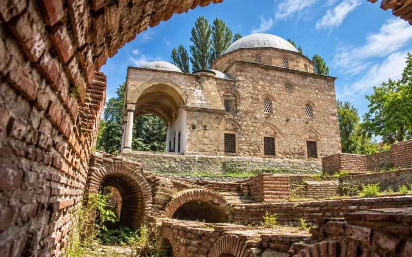 Поглед през тухлена арка към археологически останки и историческа каменна сграда с купол под синьо небе