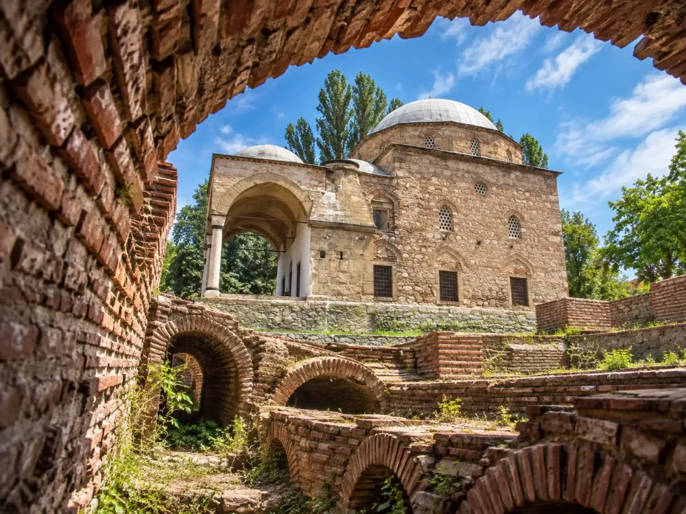 Поглед през тухлена арка към археологически останки и историческа каменна сграда с купол под синьо небе