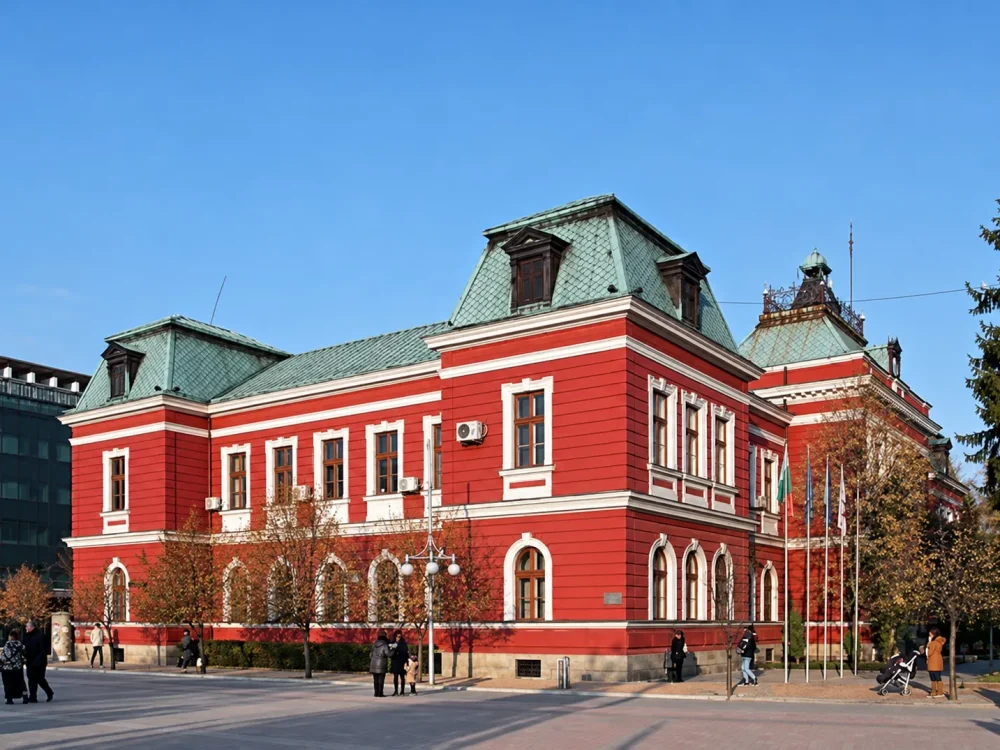 Червена сграда с бели декоративни рамки и зелен покрив край градски площад.